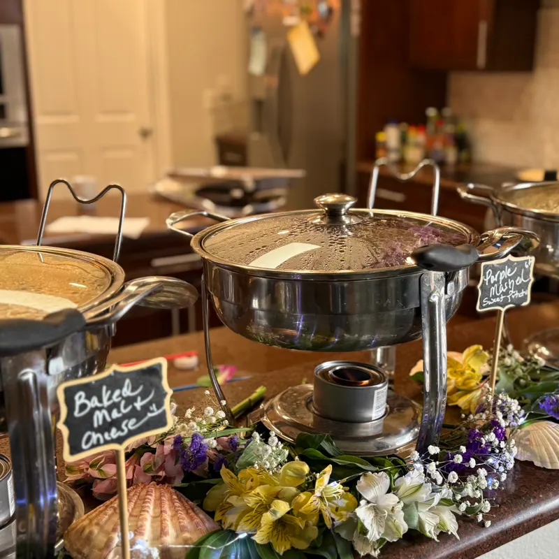 Catered buffet with chafing dishes of baked mac and cheese and purple mashed sweet potatoes, decorated with fresh flowers